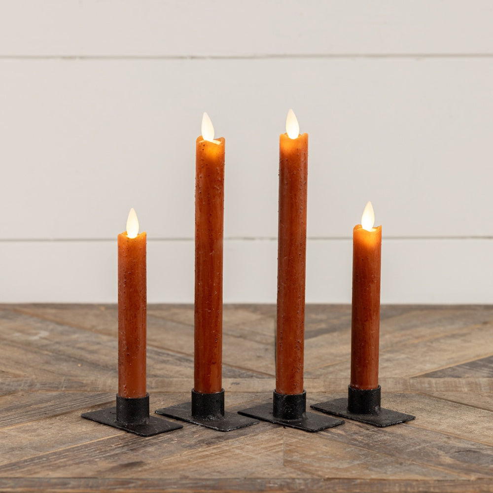 Four lit bronze-colored taper candles in black metal stands arranged on a rustic wooden surface.