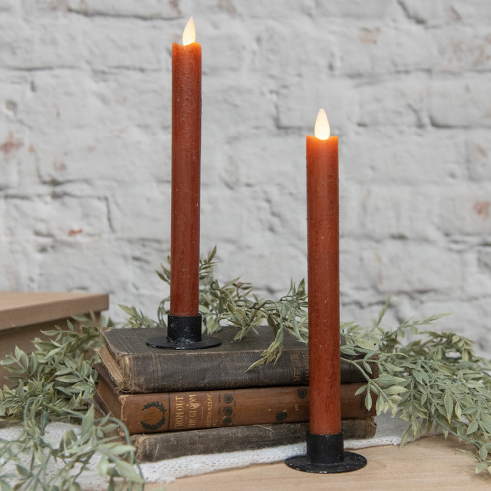 Two lit bronze taper LED candles in black holders displayed with old books and greenery against a white brick wall.