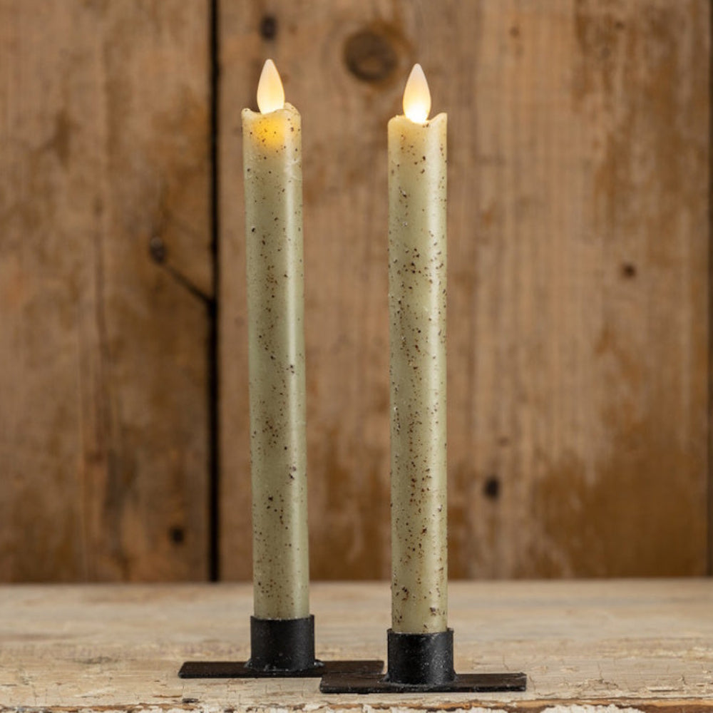 Two glowing speckled sage green taper candles in black metal bases standing on a rustic wooden surface.