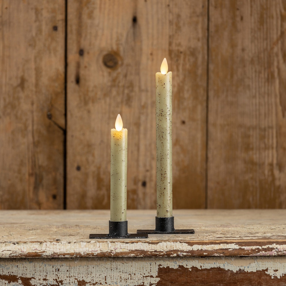 Two speckled light green taper candles in black holders glowing against a wooden wall background.