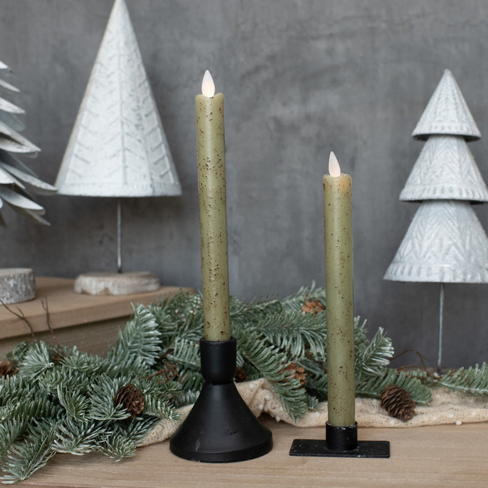 Two lit sage green taper candles in black holders with pine branches and white tree decorations behind them.