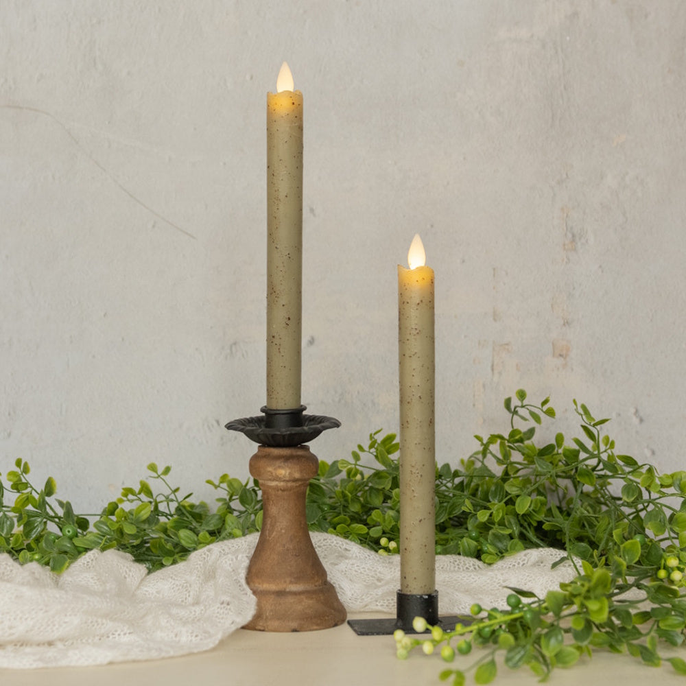 Two lit specked green taper LED candles displayed with greenery, one in a wooden holder and one on a metal stand.