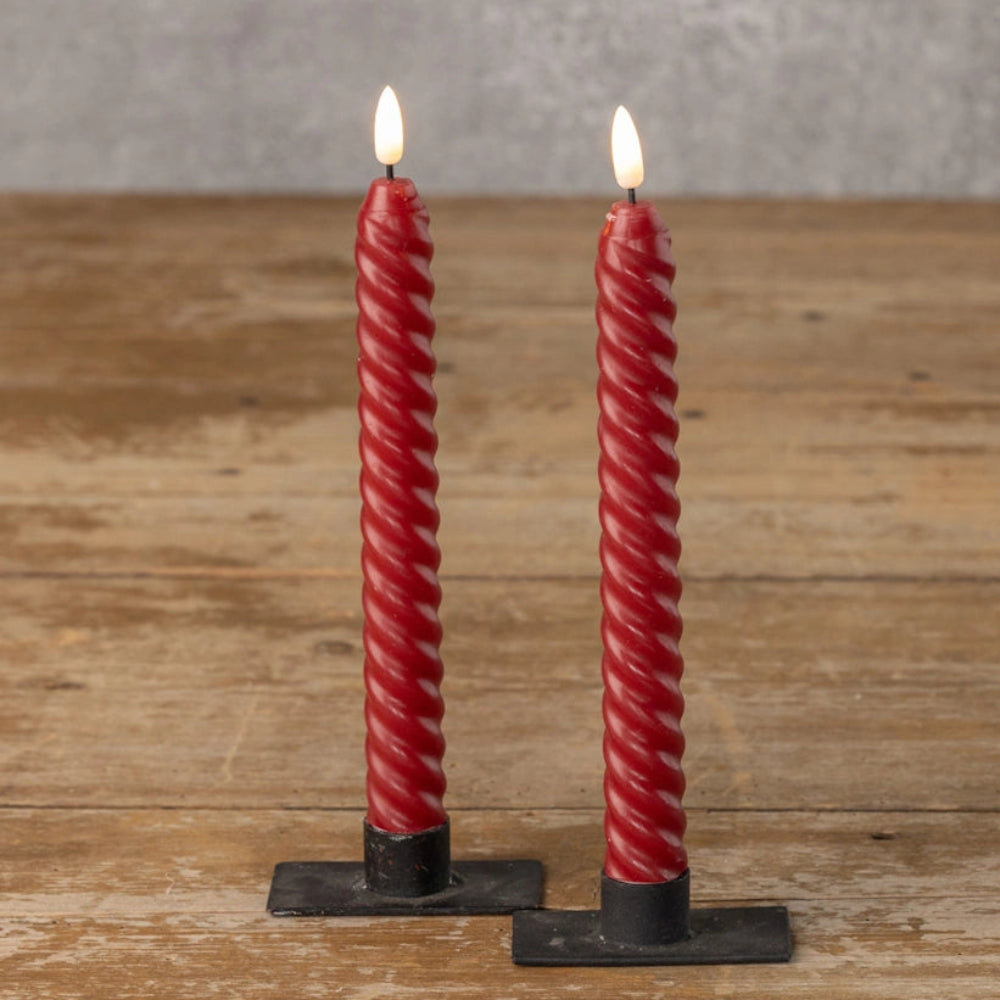 Pair of red twisted taper candles with glowing flames in black bases on a wooden table.