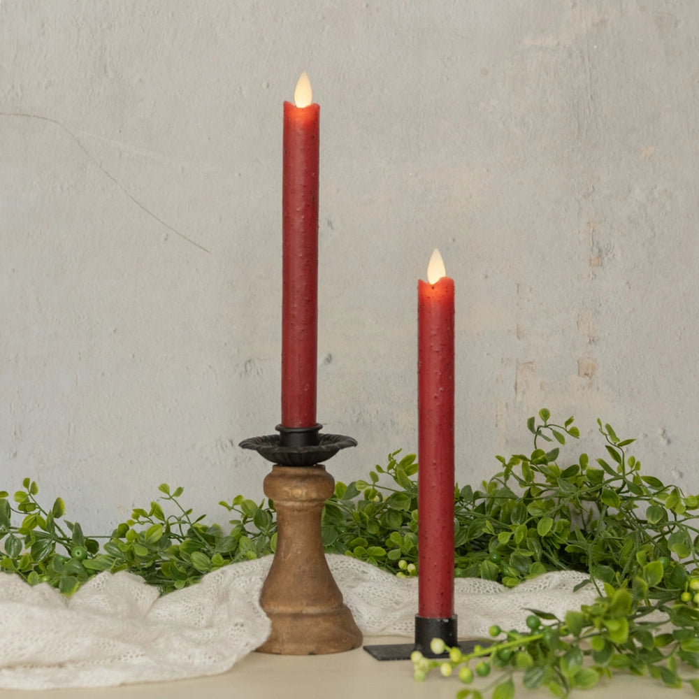 Two red flameless taper candles glowing in holders, surrounded by greenery and white fabric.