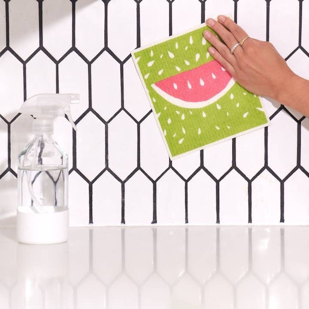 A person wipes a tiled wall with a watermelon-patterned Swedish dishcloth next to a spray bottle.