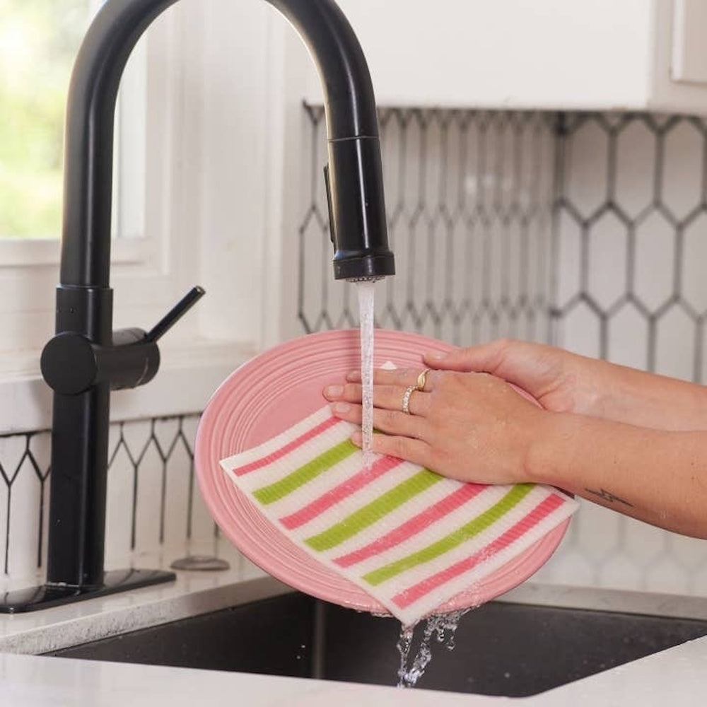 Hands washing a pink plate under a running black faucet using a striped reusable dishcloth.