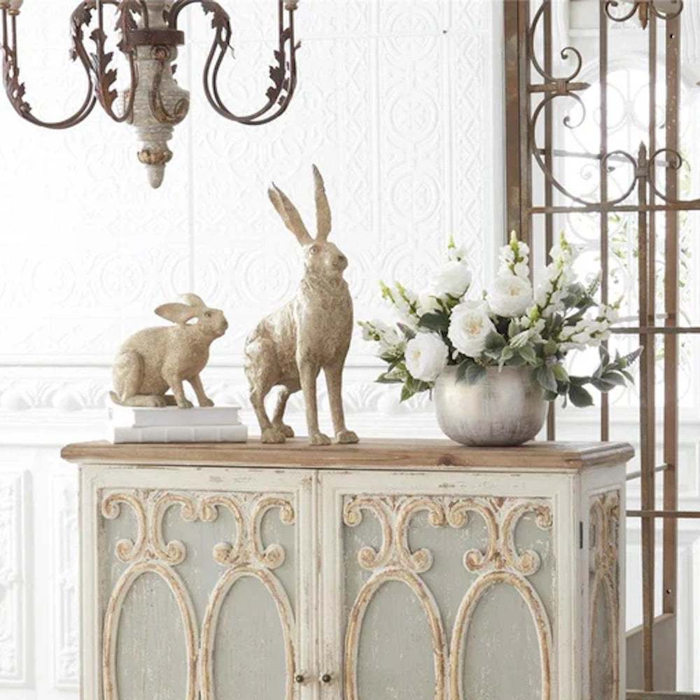 Two rustic rabbit figurines and a vase of white flowers displayed on top of a distressed vintage cabinet.