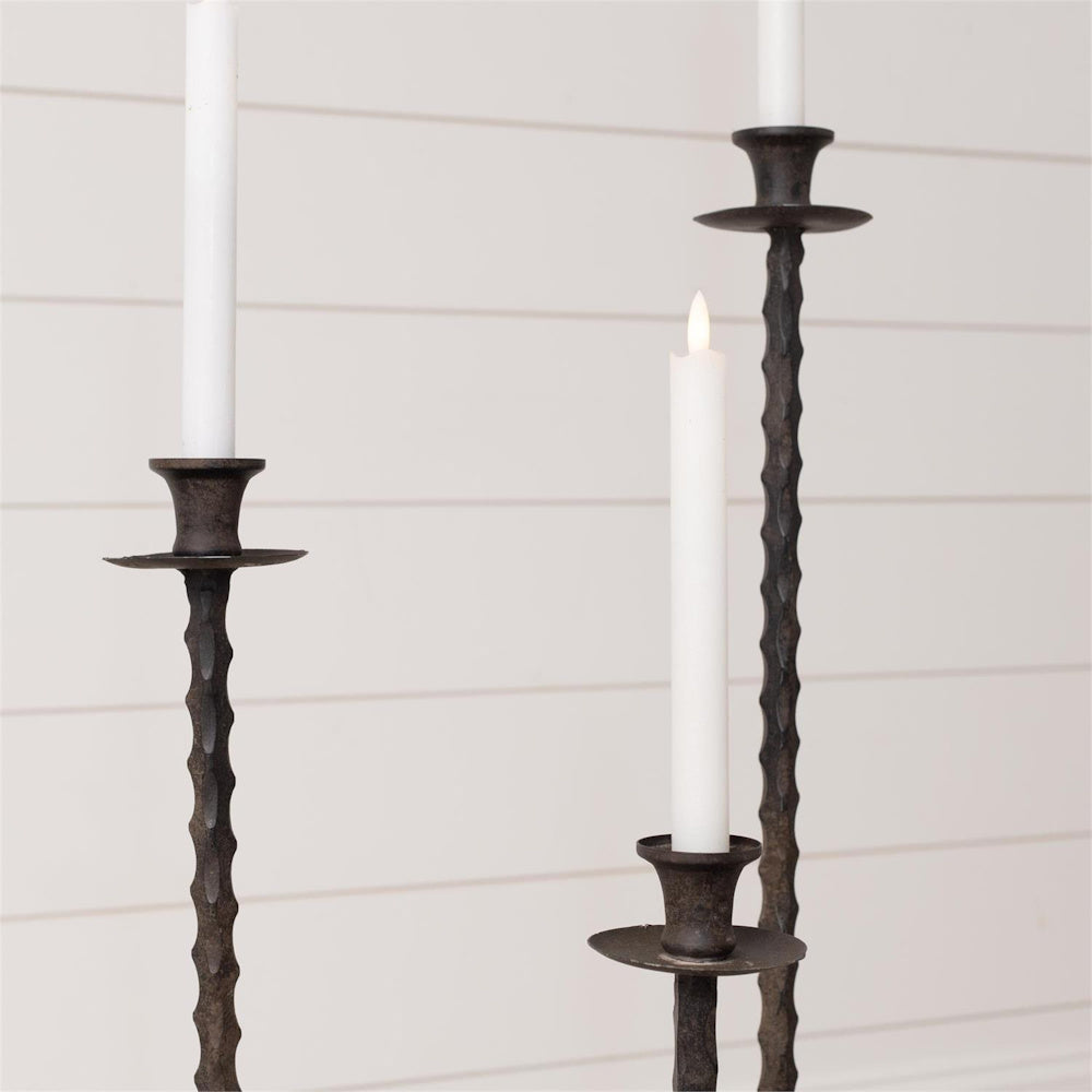 Set of 3 rustic hammered metal floor taper candle holders with white candles against a white shiplap wall.