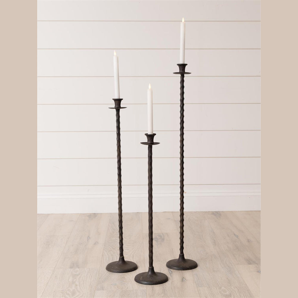 Set of 3 rustic hammered metal floor taper candle holders in varying heights with white candles on a light wood floor.