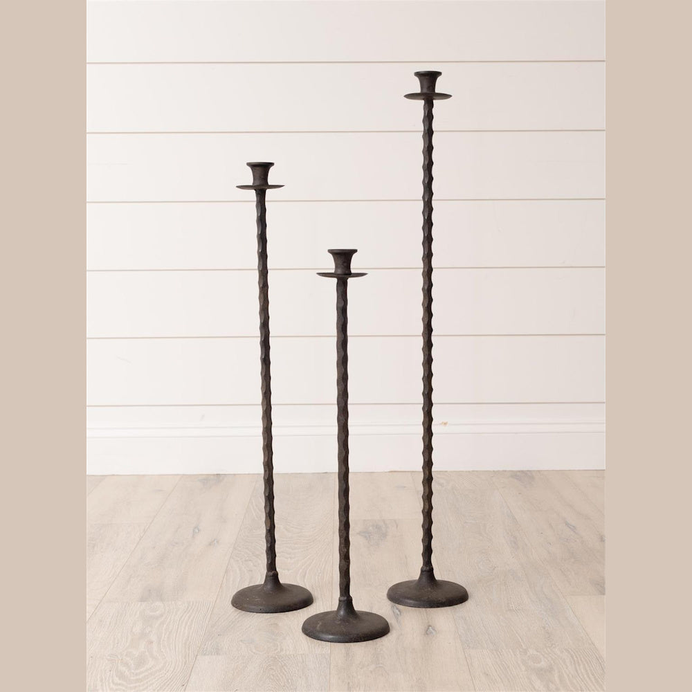 Set of 3 rustic hand-forged bronze floor taper candle holders in staggered heights against a white shiplap wall.