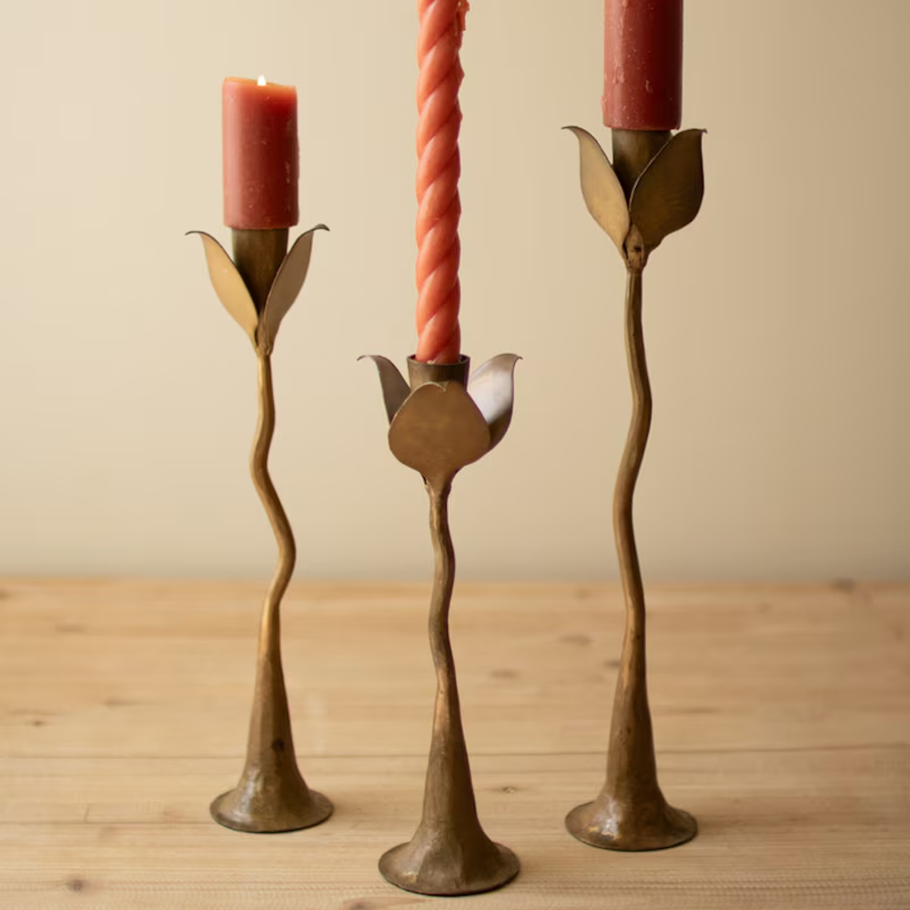 Set of three brass forged iron taper candle holders with leaf-shaped cups resting on a wooden surface.