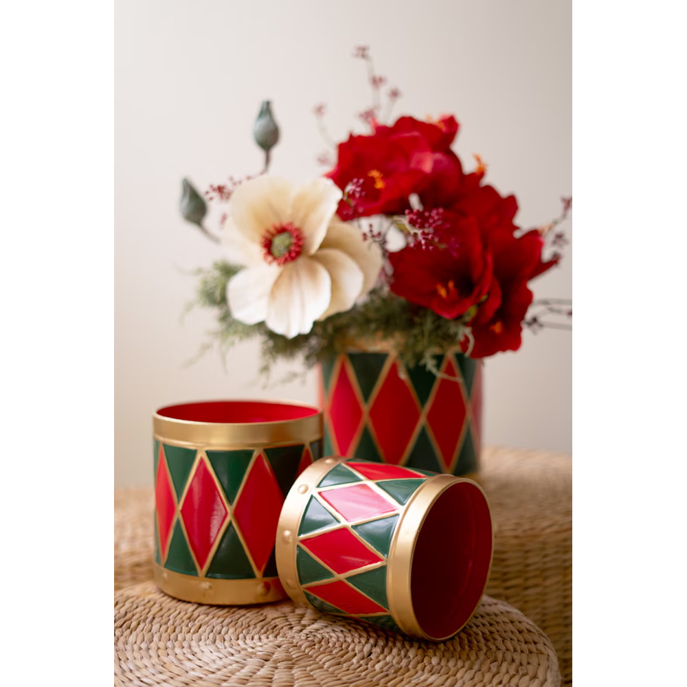 Vertical close-up of three harlequin drum planters on woven stools, one filled with red and white flowers.