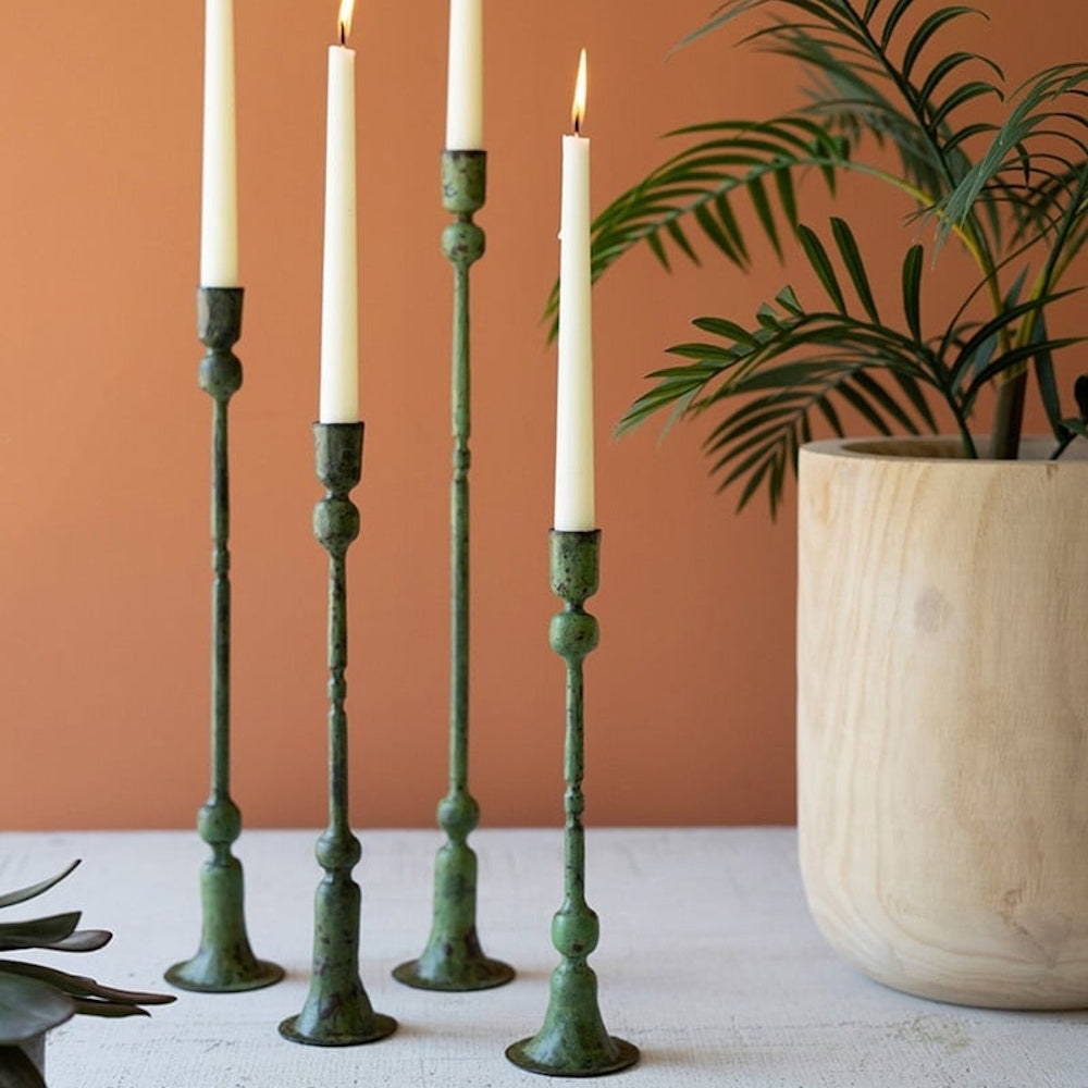 Four green patina taper candle holders with lit white candles stand next to a potted palm plant.