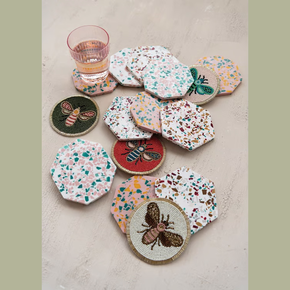 Hand-beaded insect coasters and colorful terrazzo hexagonal coasters scattered on a neutral tabletop.