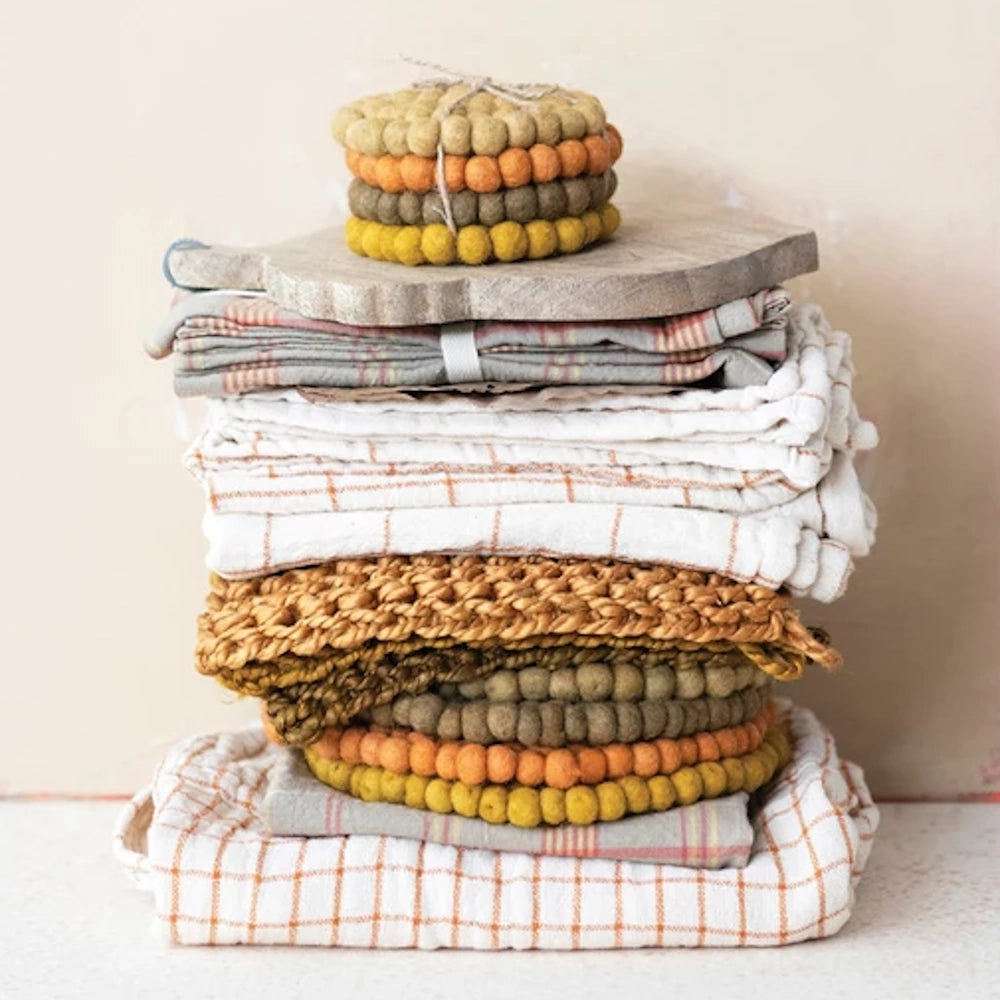Four colorful wool felt ball coasters tied with string, resting on a stack of folded linens.