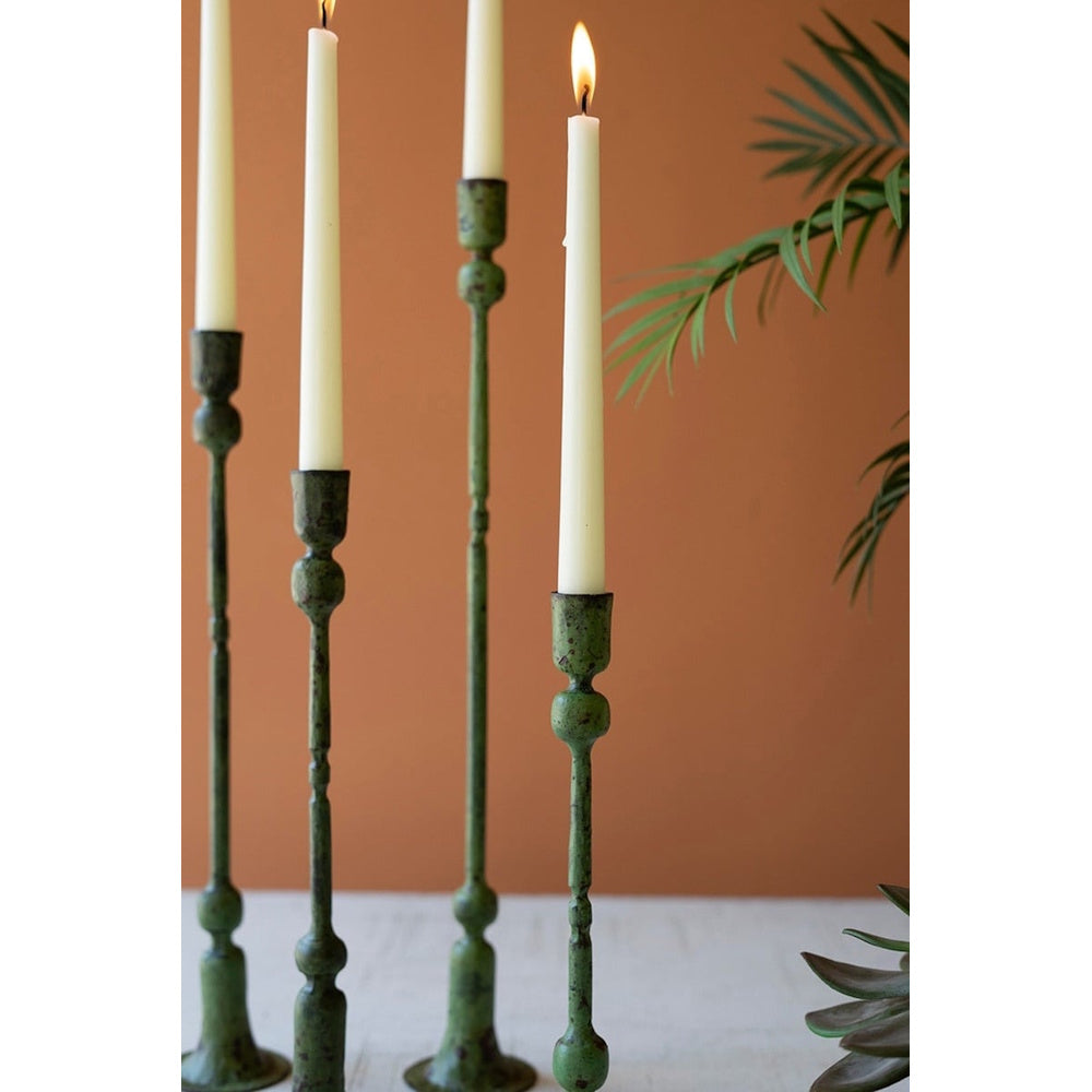 Four tall, green patina forged iron taper candle holders with lit white candles against a warm brown wall.