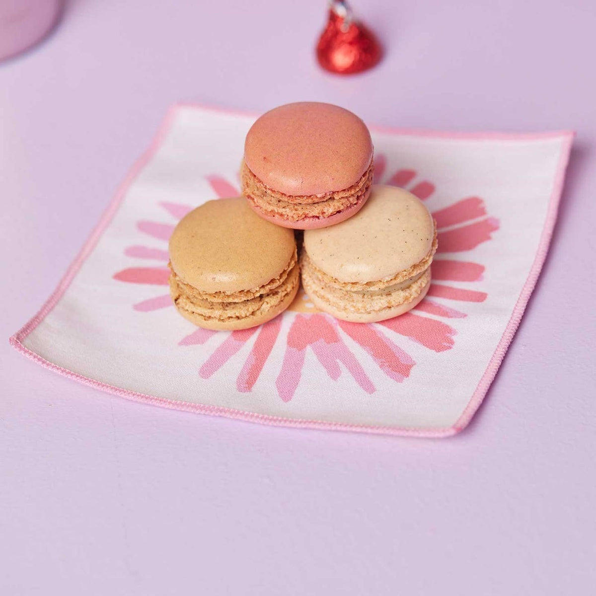 Three French macarons stacked on a white cocktail napkin with a pink daisy print against a lilac background.