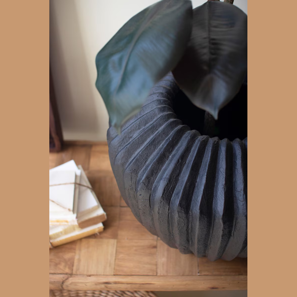 Black fluted stone composite urn planter with a matte finish, housing a large green leaf plant on a wooden surface.