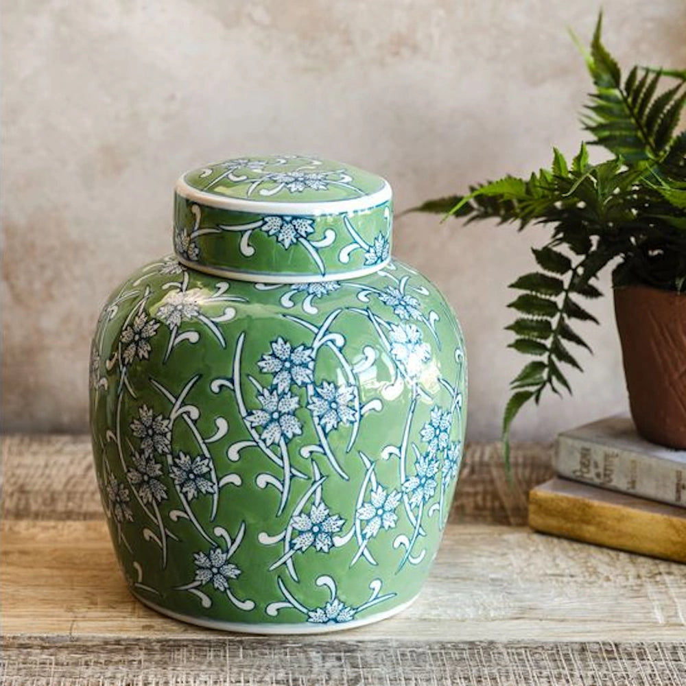 Small green and blue floral ginger jar with lid on a wooden table next to a potted fern and books.