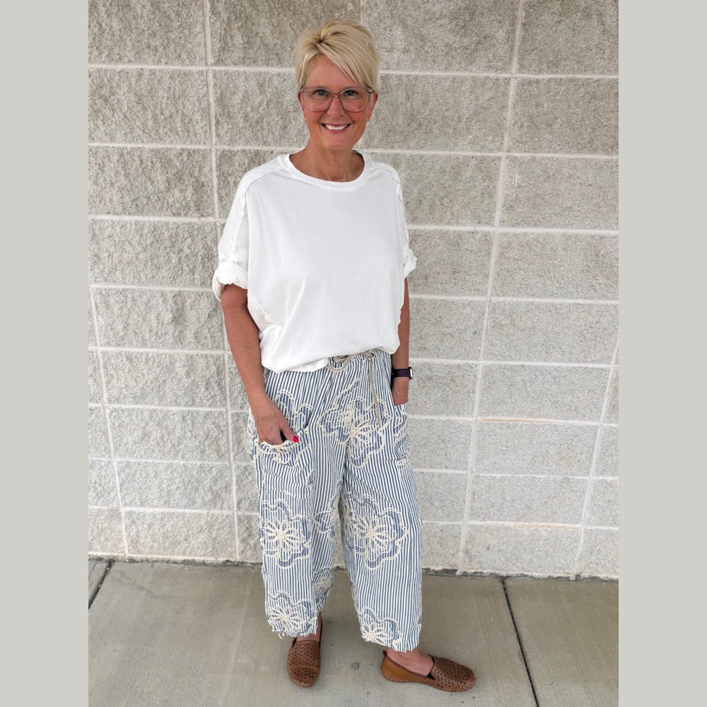 Smiling woman in a white oversized t-shirt and blue striped floral pants standing against a gray brick wall.