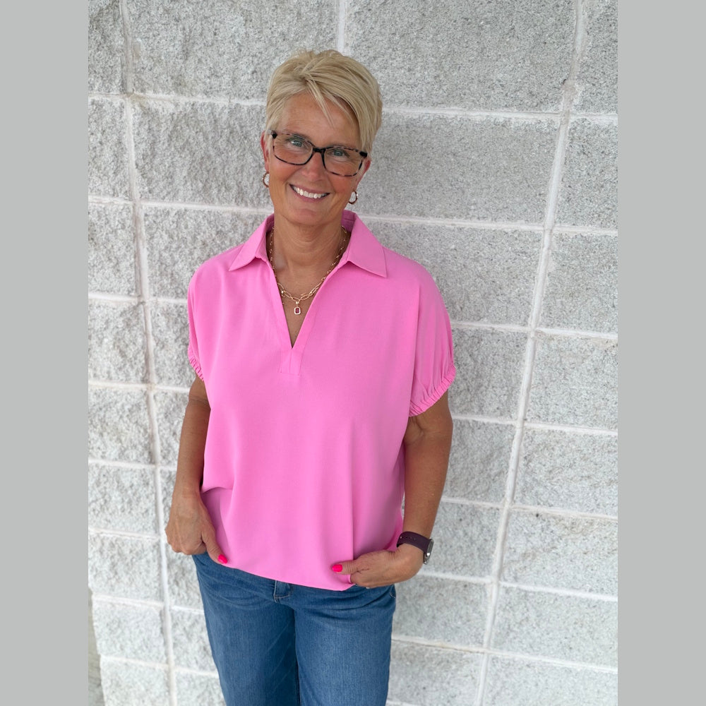 Smiling woman in a pink short-sleeve split-collar top and blue jeans, posing against a light gray brick wall.