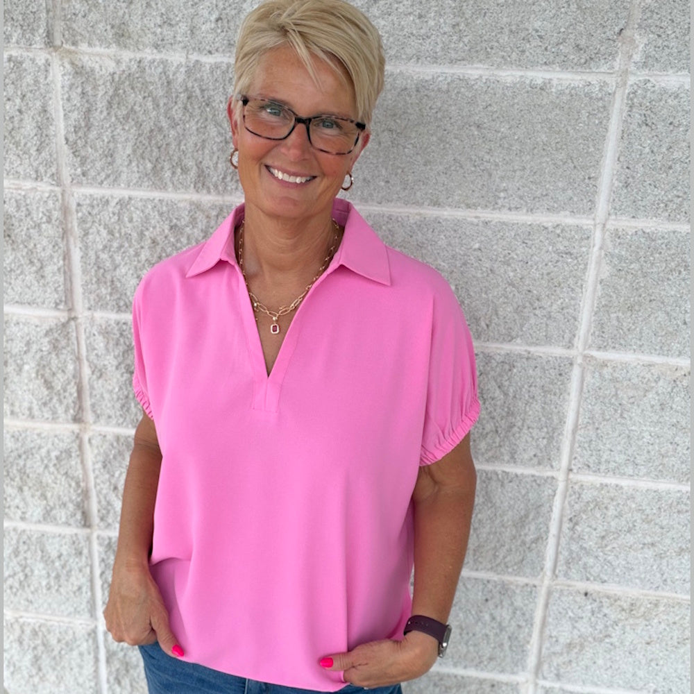 A smiling woman with blonde hair wearing a pink short sleeve split collar top and glasses against a brick wall.