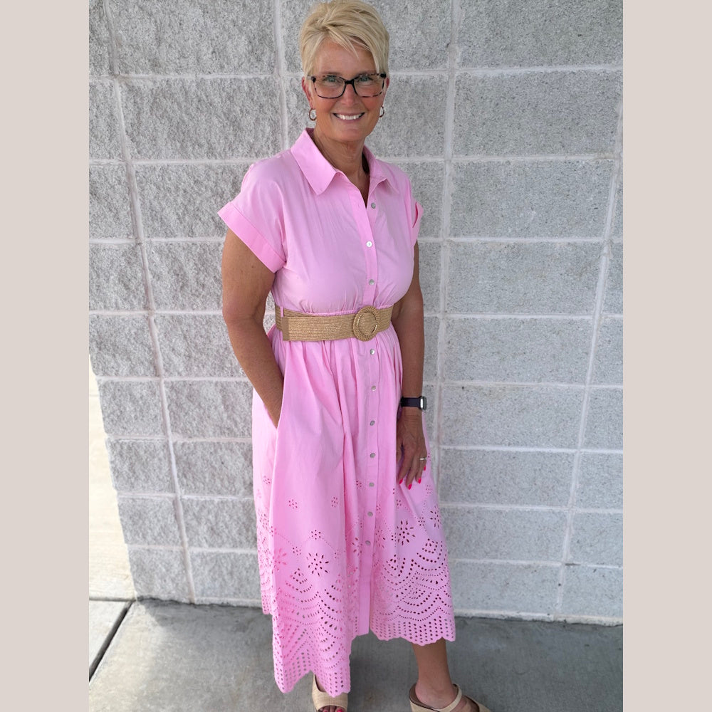 Woman in a pink short-sleeve midi shirtdress with eyelet hem, straw belt, and glasses posing against a stone wall.