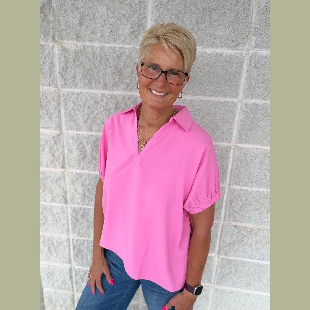 A woman with short blonde hair and glasses wearing a pink short-sleeve split-collar top and blue jeans.
