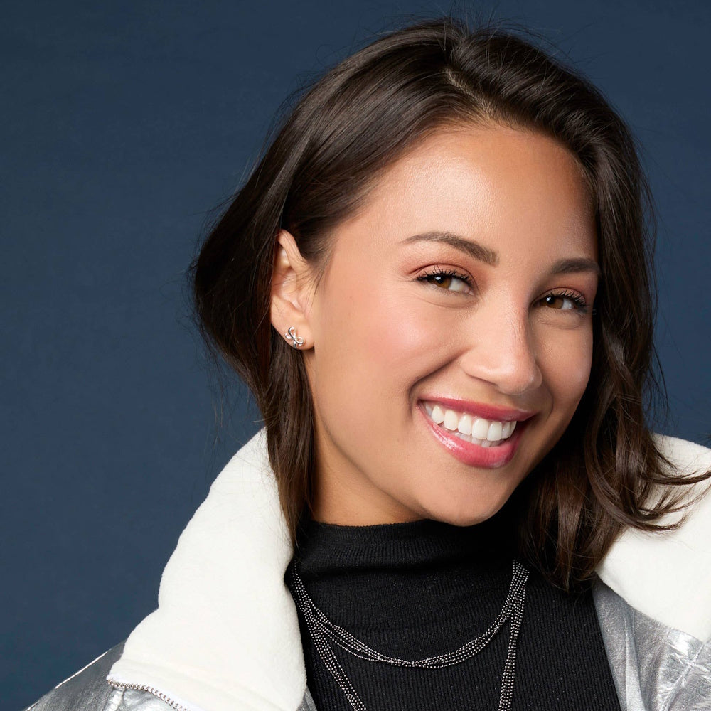 Smiling woman wearing a silver bow stud earring and a silver jacket against a dark blue background.