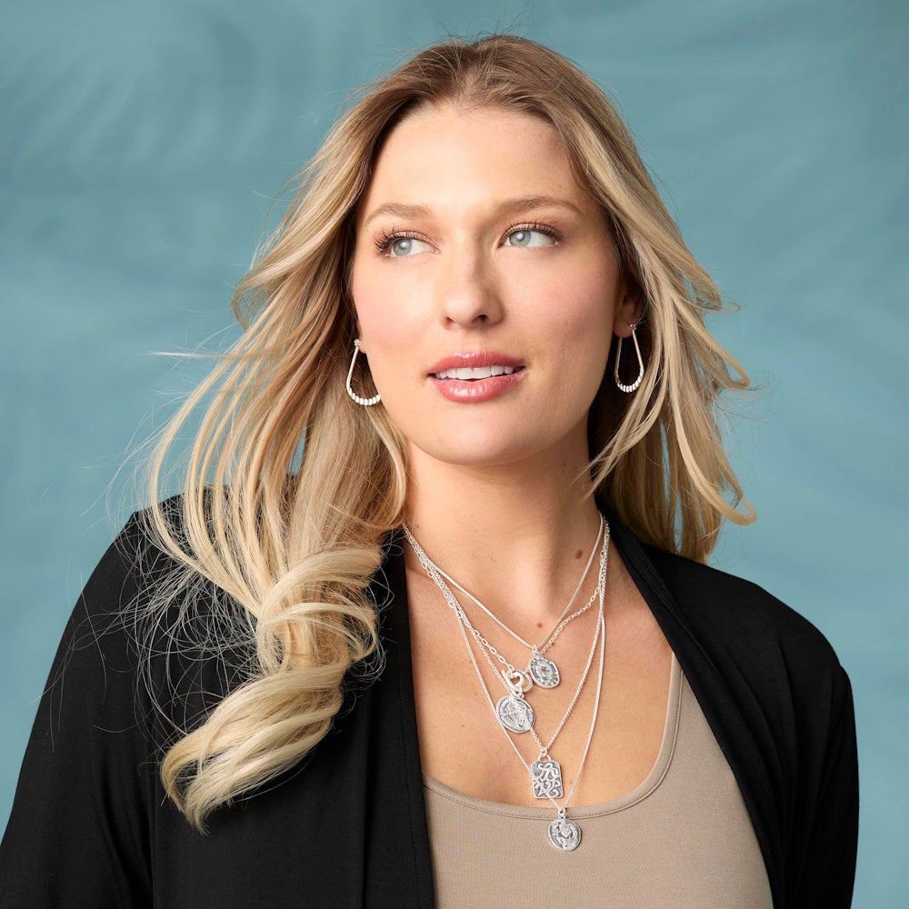 Woman wearing silver crinkle teardrop dangle earrings and layered pendant necklaces against a blue background.