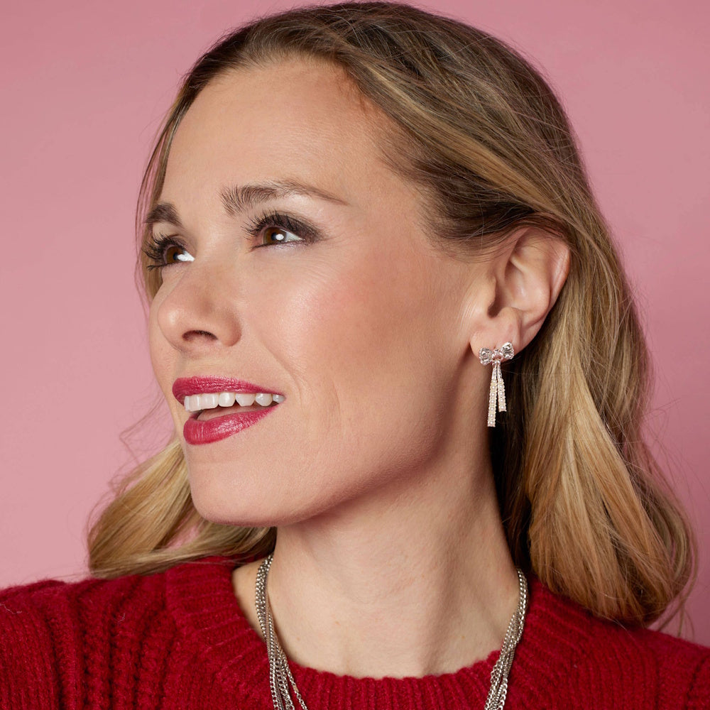 Woman smiling in red sweater wearing a silver bow dangle earring with a pink background.
