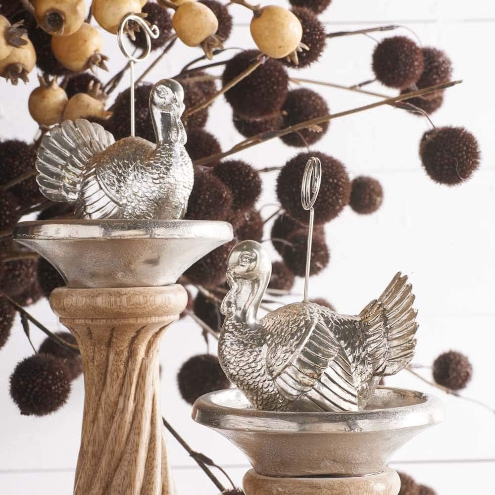Silver turkey-shaped place card holders with wire clips set on pedestals with botanical decor.