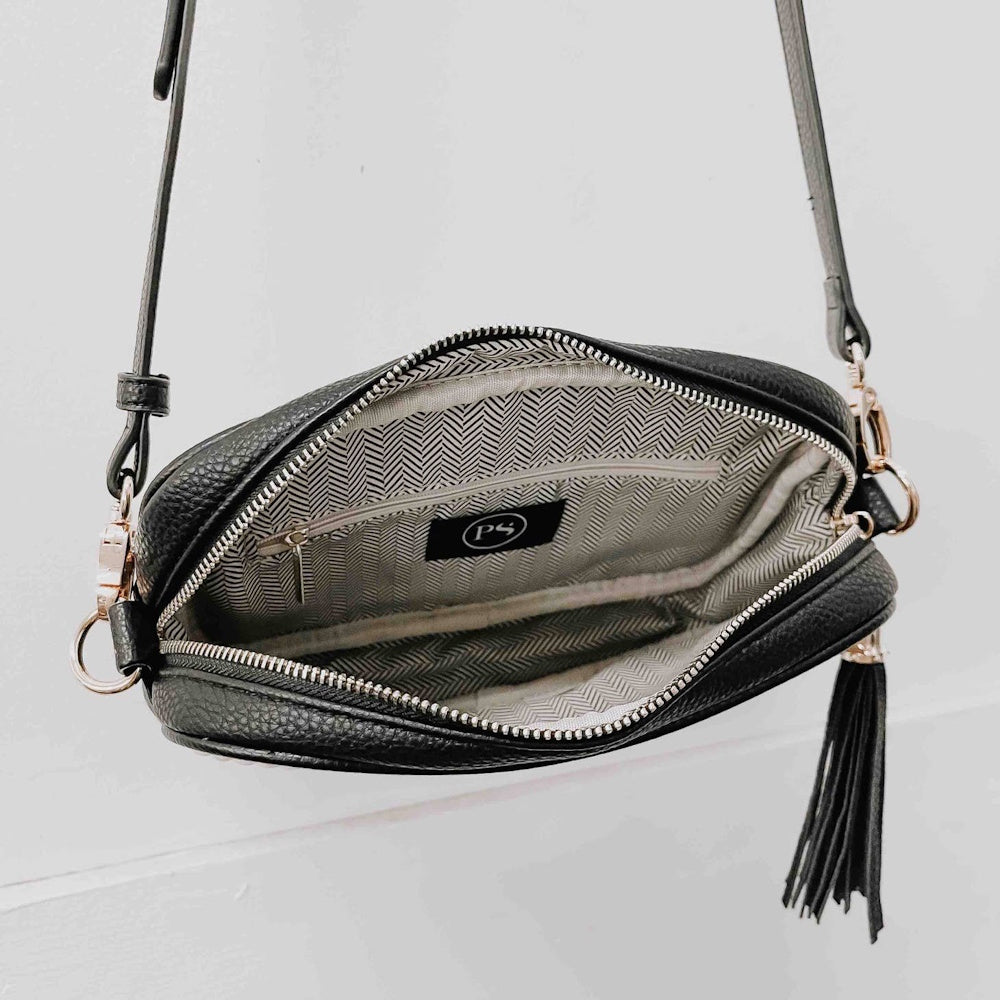 Open black leather crossbody bag showing a patterned lining, interior zip pocket, and hanging tassel.