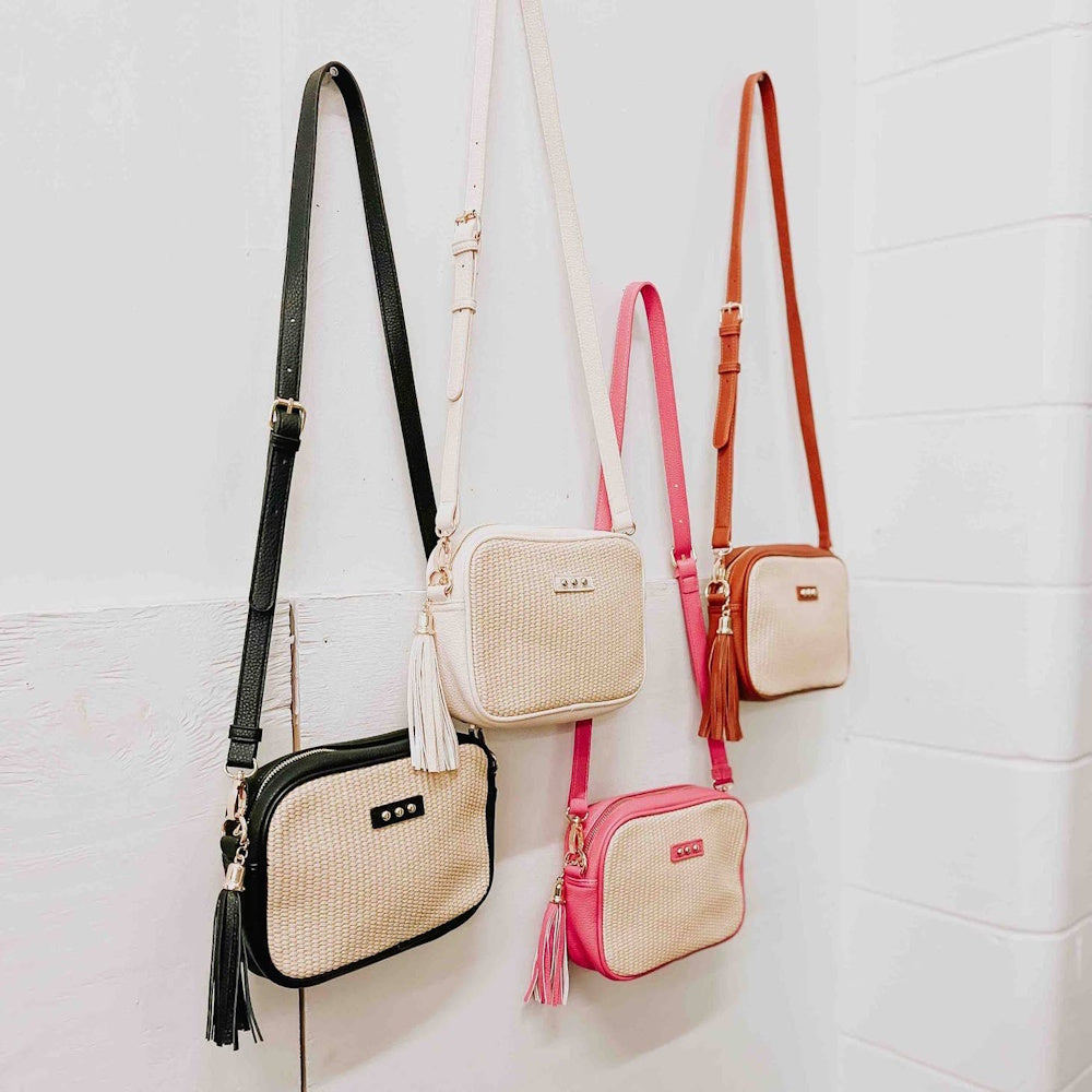 Four woven straw camera bags with tassels in black, cream, pink, and brown hanging on a white wall.