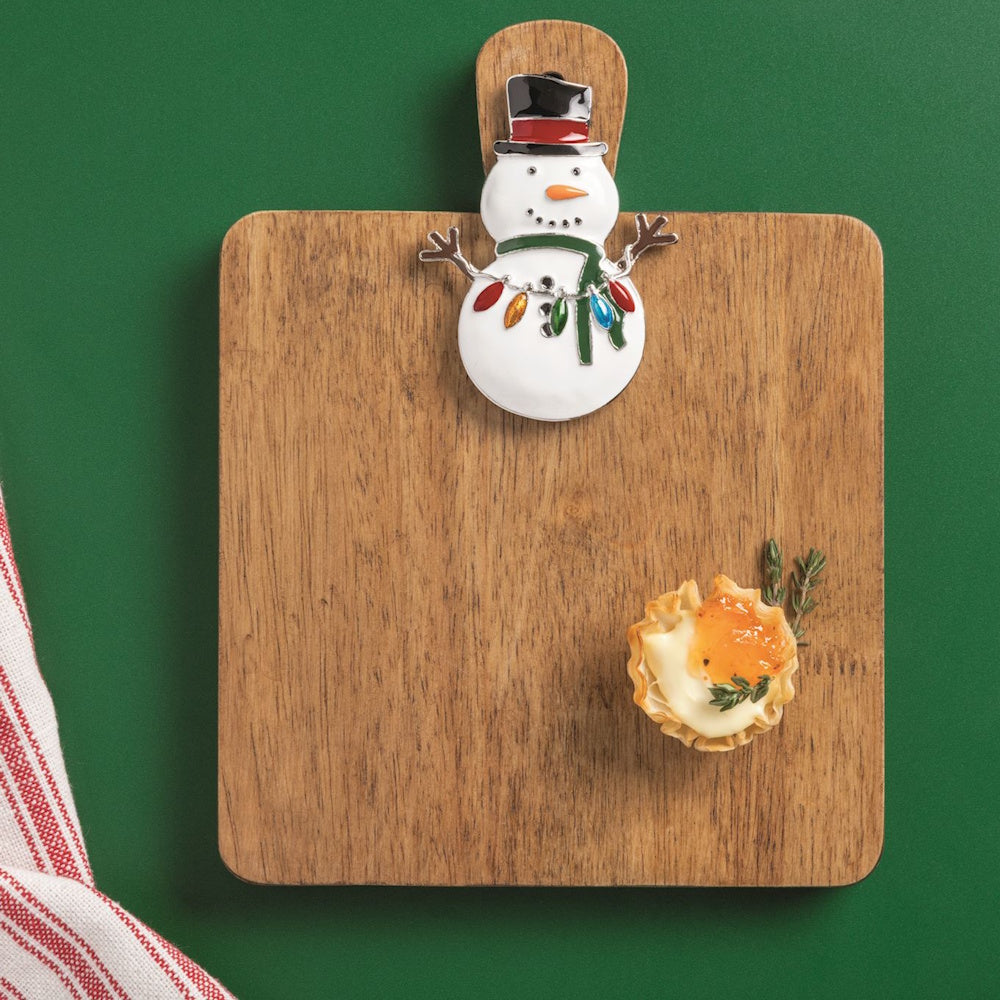 Wooden charcuterie cutting board featuring a decorative Christmas snowman charm on a green background.