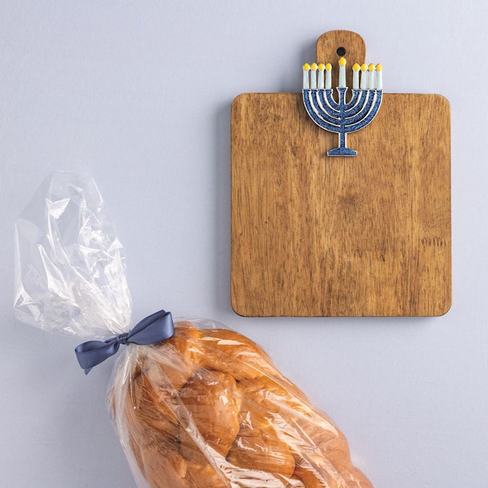 Wooden cutting board with a blue Hanukkah menorah charm next to a wrapped challah bread loaf.