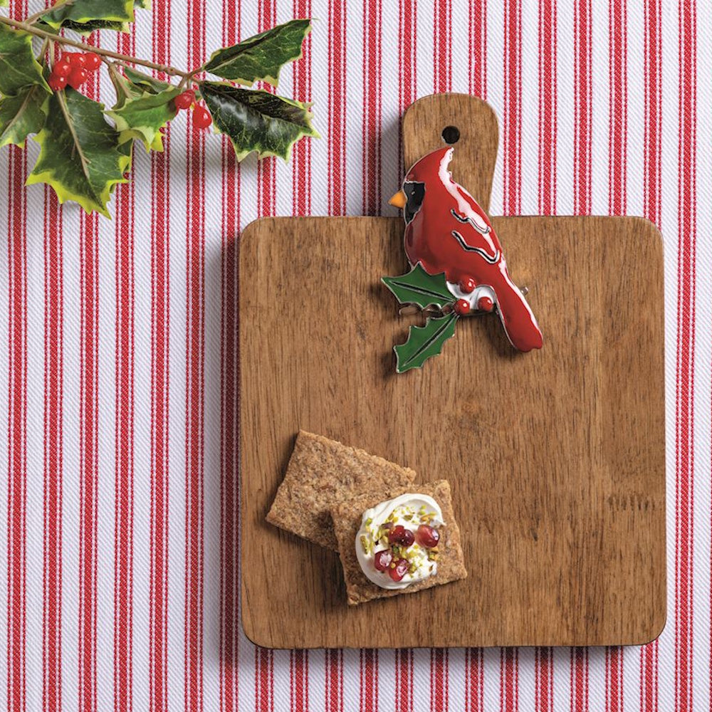 Wooden serving board with a red cardinal charm, crackers, and holiday decor on red-and-white stripes.