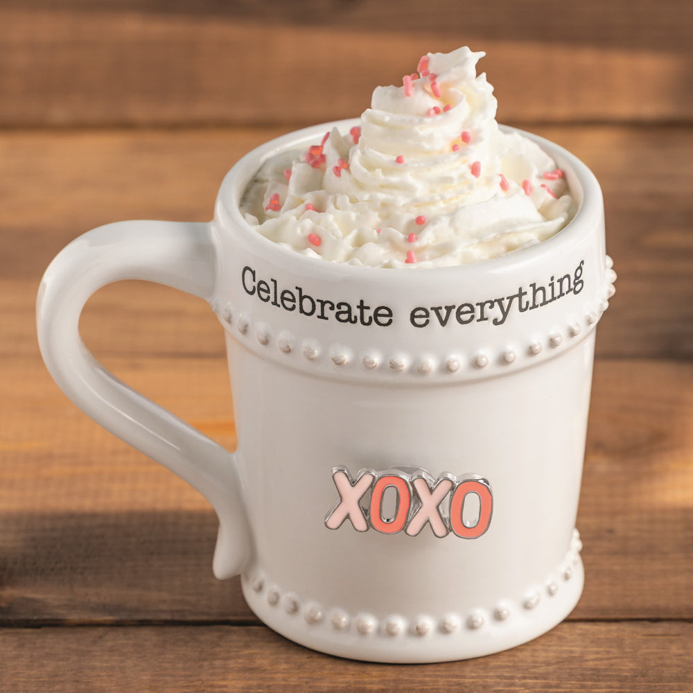White ceramic mug with "Celebrate everything" text, pink XOXO charm, and whipped cream with sprinkles on a wood background.