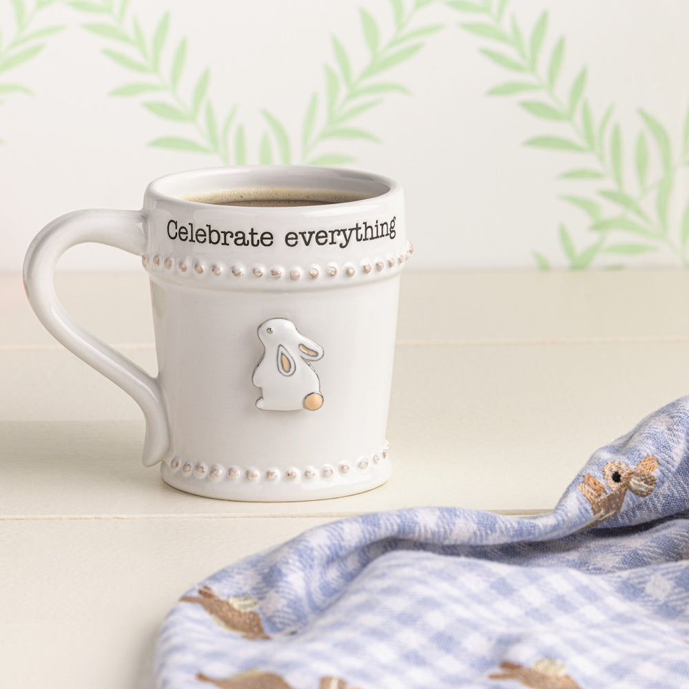 White ceramic mug with "Celebrate everything" text, a raised bunny charm, and beaded trim on a white table.
