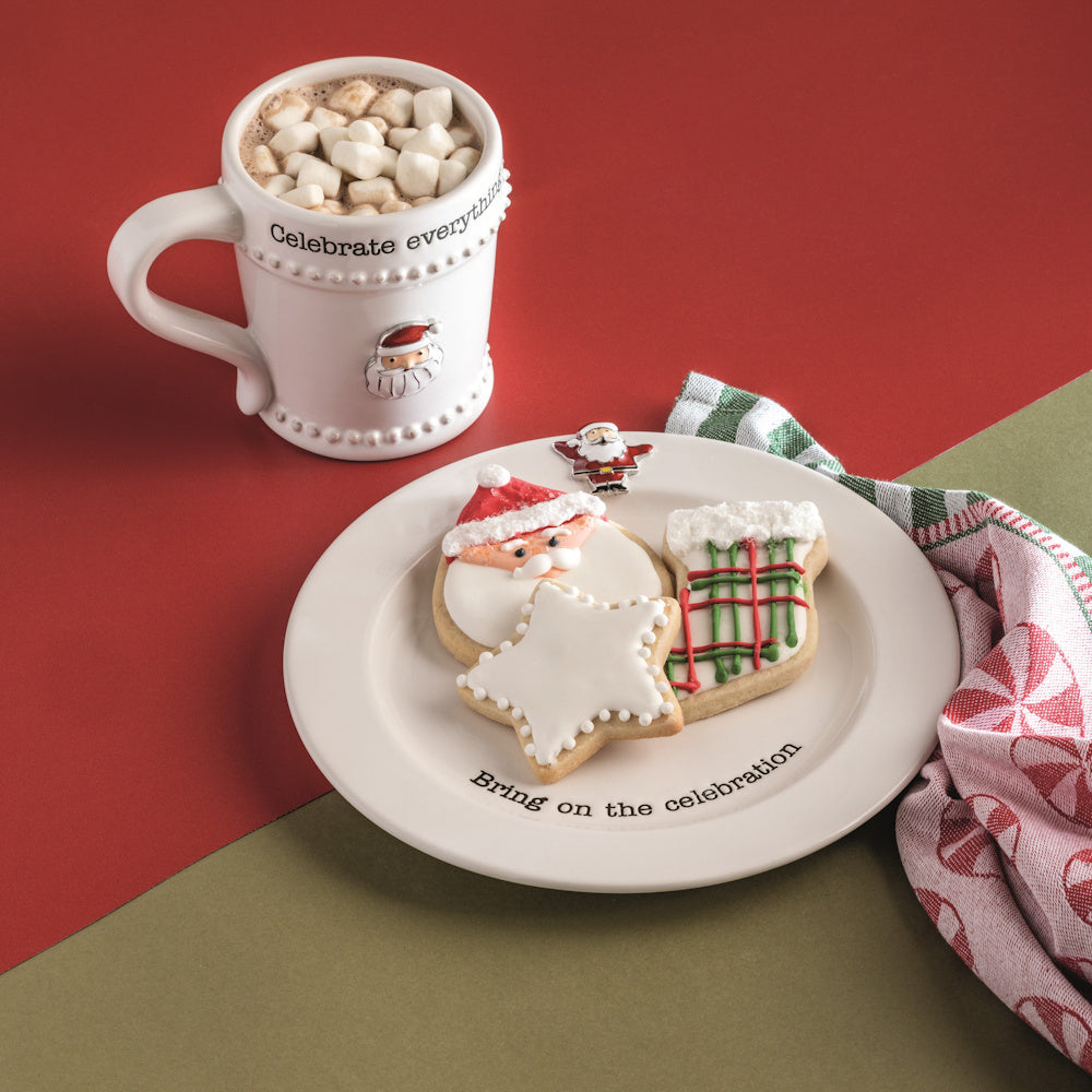 Christmas mug with marshmallows and Santa cookies on a holiday plate. White ceramic dishware on a red and green background.