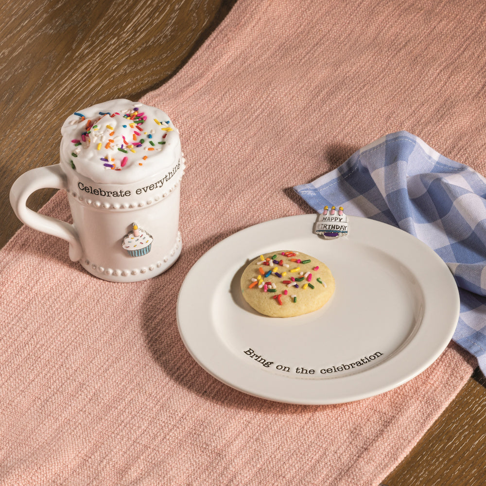 Celebrate Everything mug and matching plate with birthday charms, sprinkles, and a cookie on a pink table runner.