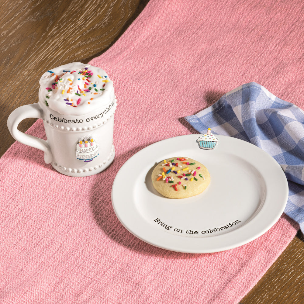 White ceramic birthday mug and plate set with a sugar cookie, sprinkles, and cake charms on a pink textured table runner.