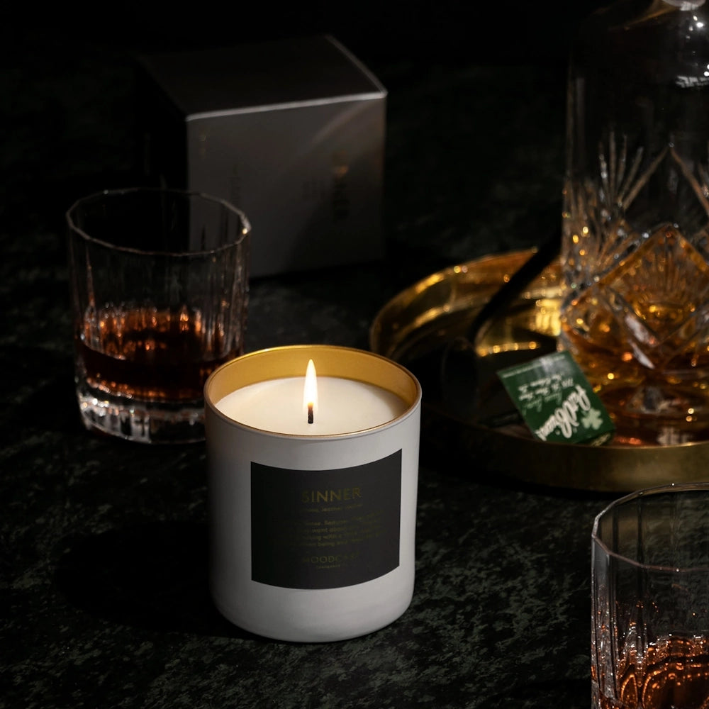 Lit Moodcast Sinner candle on a dark surface surrounded by whiskey glasses and a decanter.