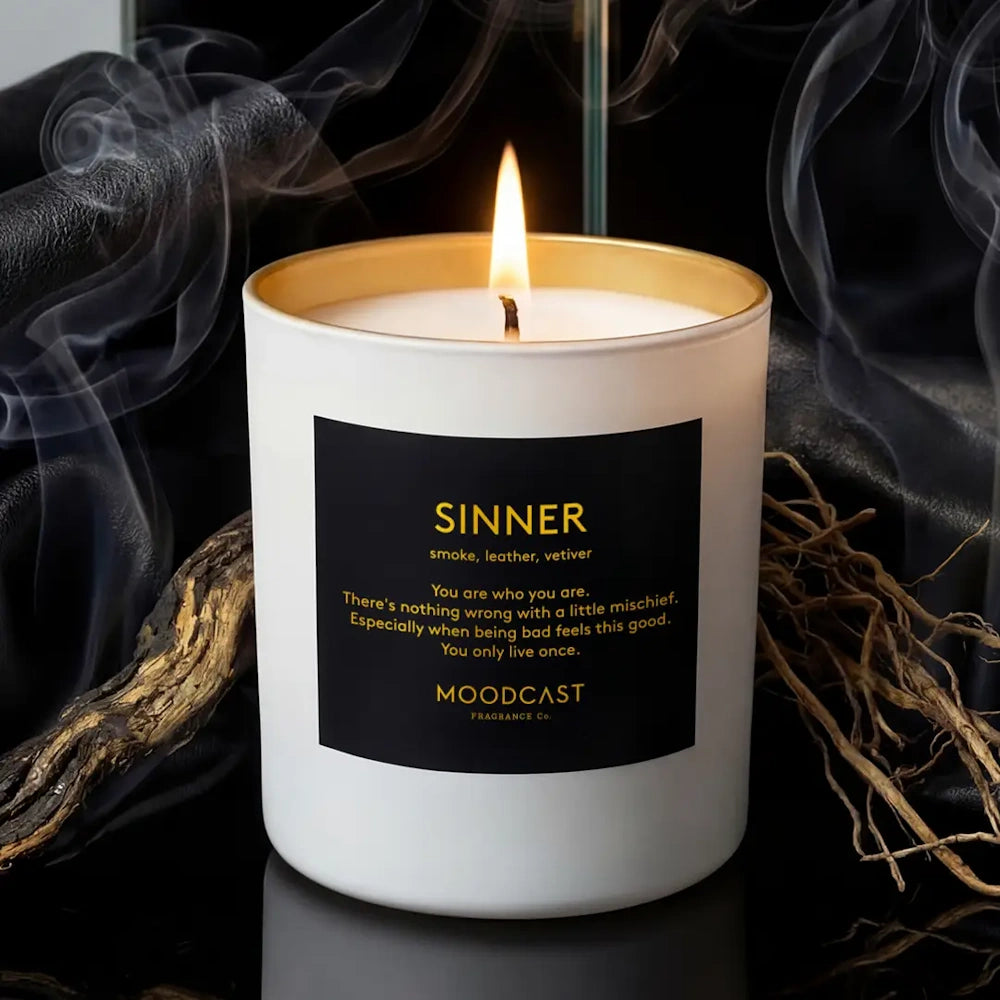 Lit Moodcast Sinner candle with a black label, surrounded by wisps of smoke, leather, and dried roots.