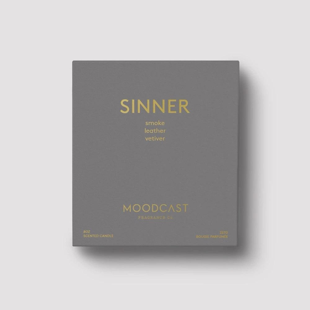 Matte dark gray Moodcast Sinner candle box packaging featuring gold metallic lettering and scent notes.