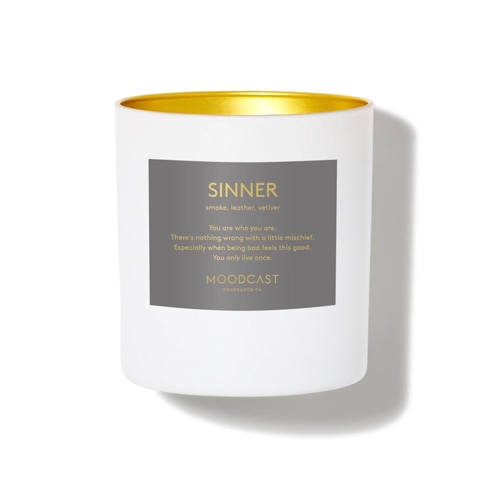 Moodcast Sinner candle in a white jar with a metallic gold interior and a dark gray rectangular label.