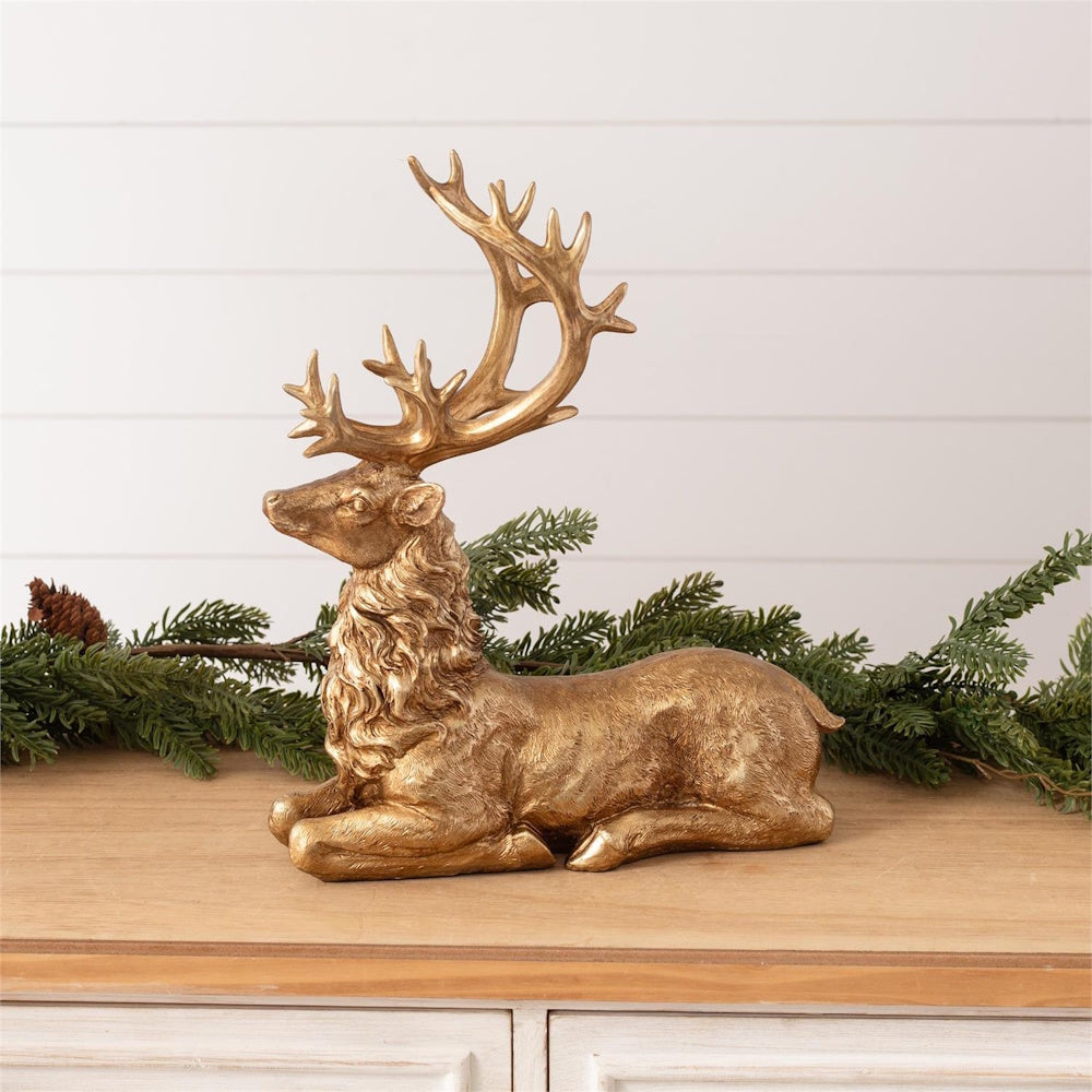 A seated gold reindeer figurine resting on a wooden surface in front of pine greenery.