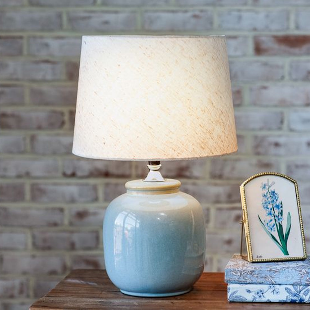 Slate blue ceramic table lamp with a linen shade on a wooden table against a white brick wall.