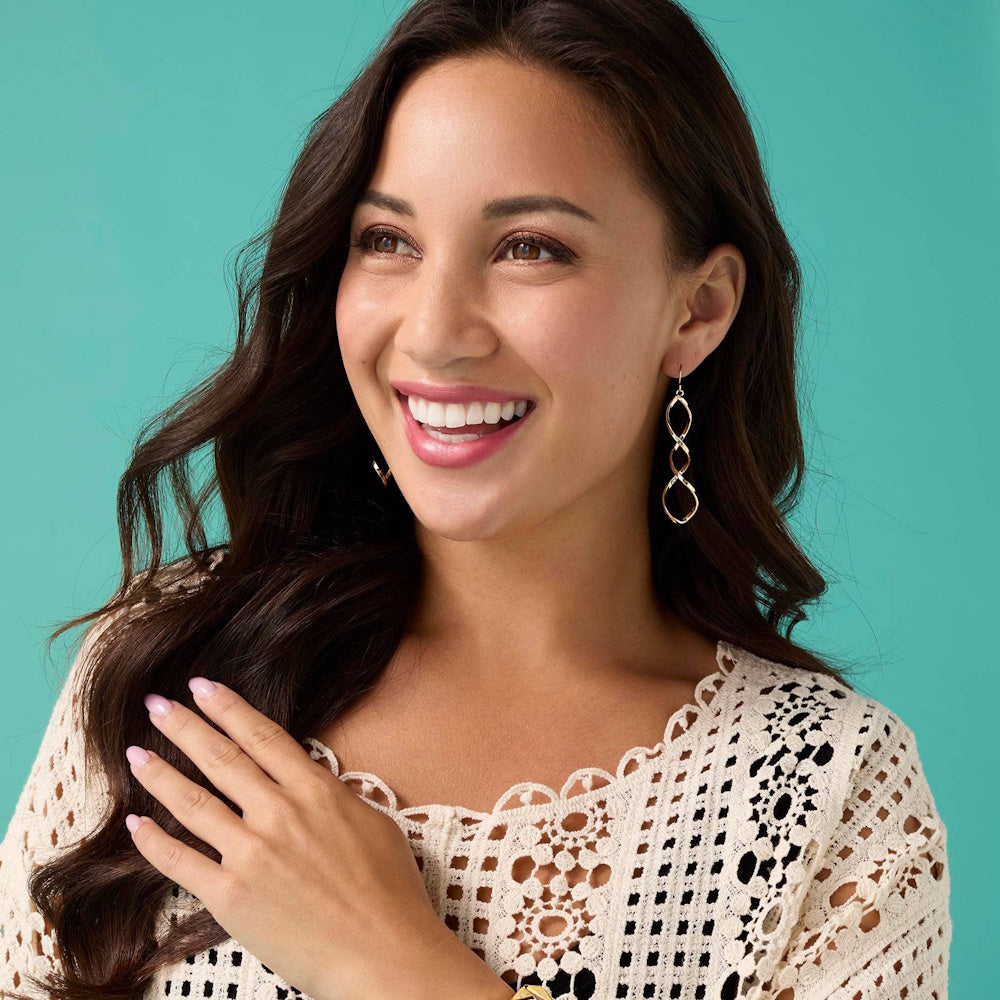 Woman smiling and wearing sleek gold twist dangle earrings and a white lace crochet top against a teal background.