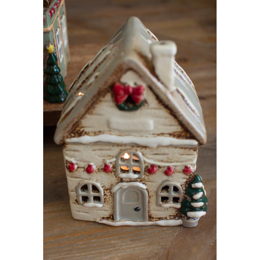 Small cream-colored ceramic Christmas village house featuring a red wreath and warm lit interior glow.